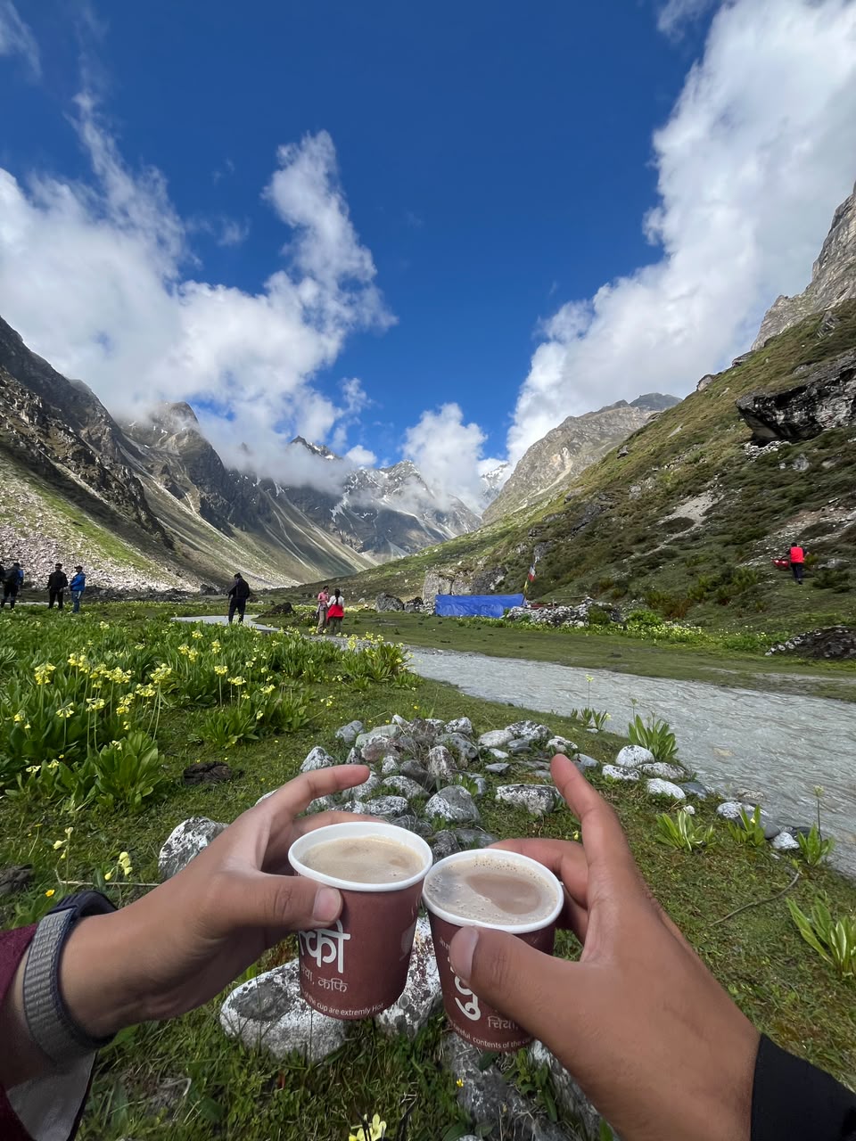 Two hands raising cups of milk tea against a view down the valley
