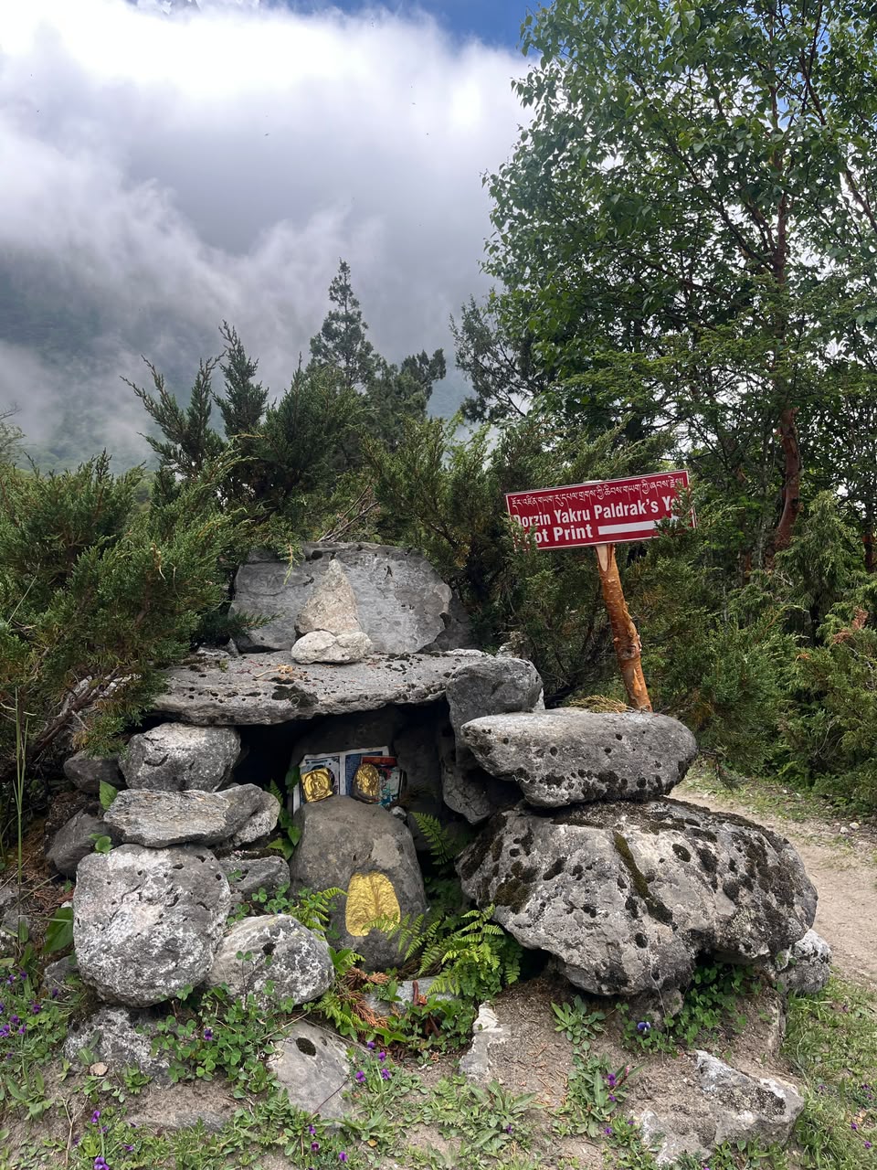 A stone shrine along the trail marking a yeti footprint, with juniper and cloud-covered peaks behind